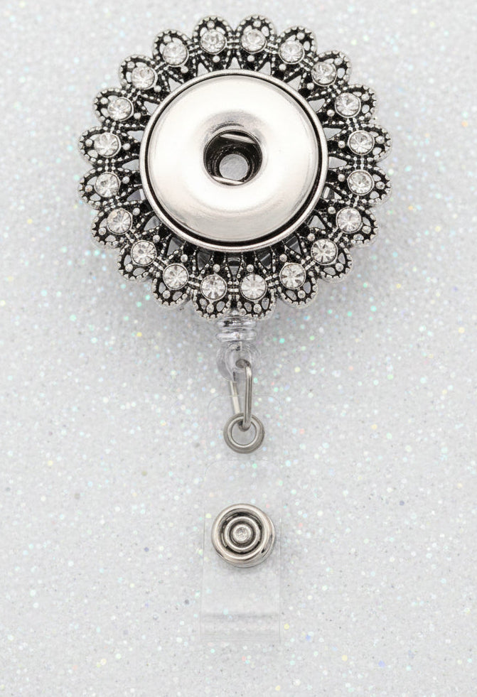A silver-toned retractable badge reel with a decorative floral-style metal top and a clear plastic clip below, shown on a white background with a black shape behind it.