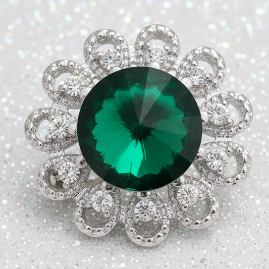 A silver-toned floral cluster snap button featuring a large round faceted green emerald-cut center stone surrounded by a halo of smaller clear stones in an openwork silver setting, shown on a plain white background.