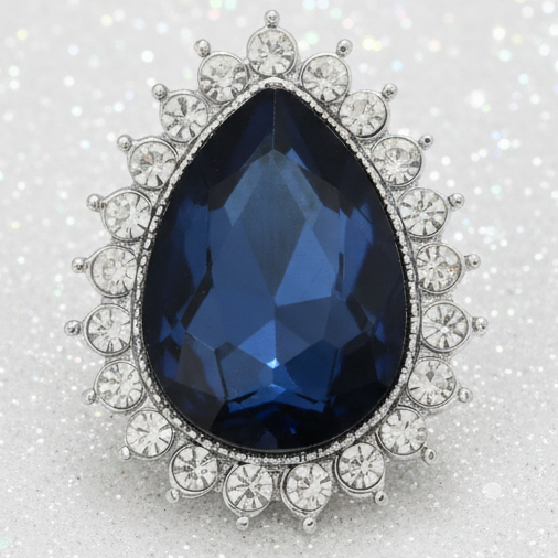 A pear-shaped blue sapphire snap set in a silver-toned halo with surrounding round diamonds, photographed on a sparkling white background.