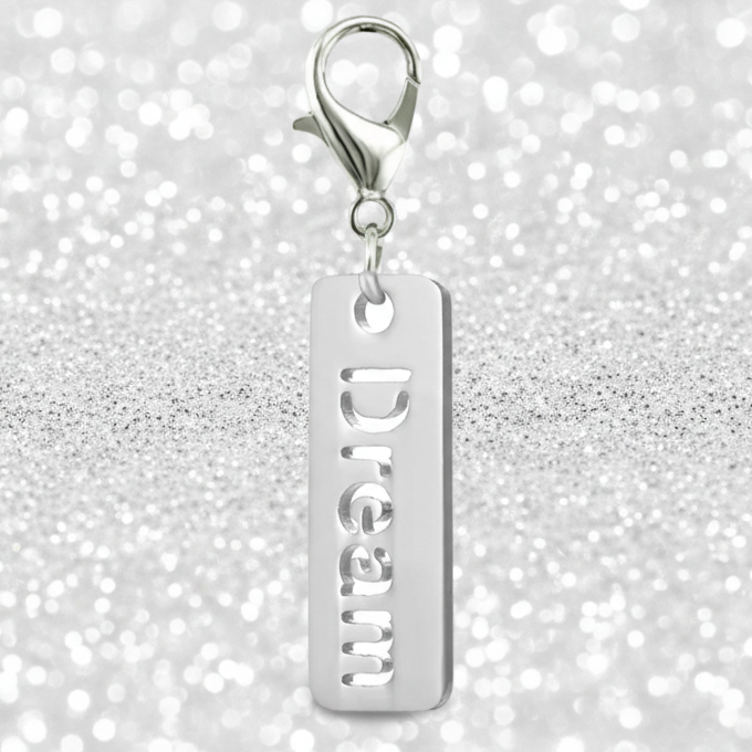 A silver rectangular charm with the word 'Dream' cut vertically, hanging from a silver lobster clasp, set against a sparkling silver glitter background.
