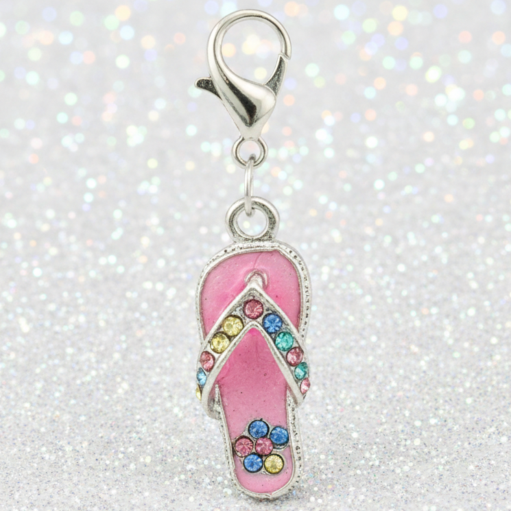 A pink flip-flop charm with a silver clasp, decorated with colorful rhinestones and a small blue flower, set against a sparkly silver glitter background.