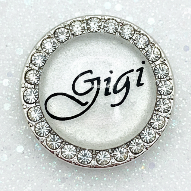 A round silver-toned snap with a border of clear rhinestones and the name 'Gigi' written in black script in the center.