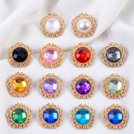 Twelve ornate gold-tone square snap buttons arranged in a 4-by-3 grid on a black background. The top two have round white faux pearls, while the remaining ten have faceted gemstones in black, pink, red, gray, yellow, purple, mint green, blue, and light blue.