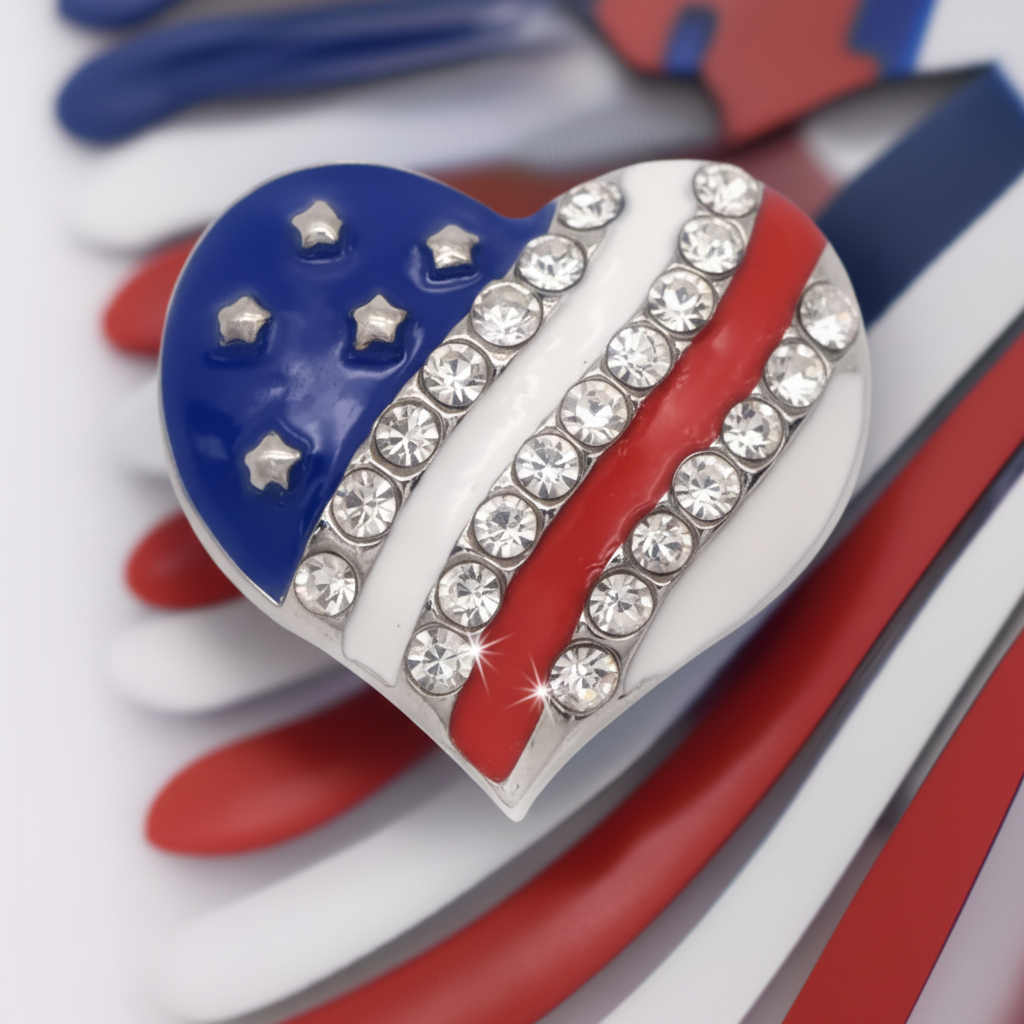A heart-shaped snap button featuring an American flag design with blue, white, and red sections, embellished with clear rhinestones along the white stripes, shown on a red, white, and blue striped background.