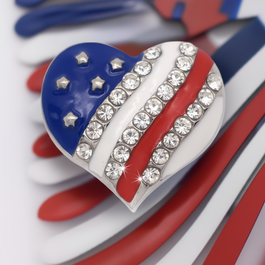 A heart-shaped snap button featuring an American flag design with blue, white, and red sections, embellished with clear rhinestones along the white stripes, shown on a red, white, and blue striped background.