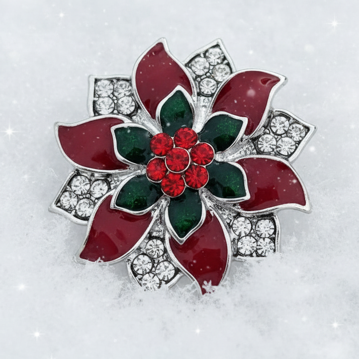 A festive poinsettia flower brooch with red enamel petals, green leaves, a red crystal center, and silver crystal-studded accents, displayed on a snowy white background with a subtle starry sparkle overlay.