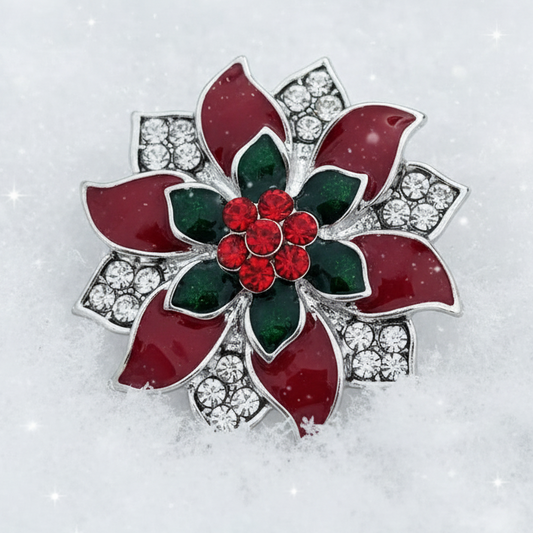 A festive poinsettia flower brooch with red enamel petals, green leaves, a red crystal center, and silver crystal-studded accents, displayed on a snowy white background with a subtle starry sparkle overlay.