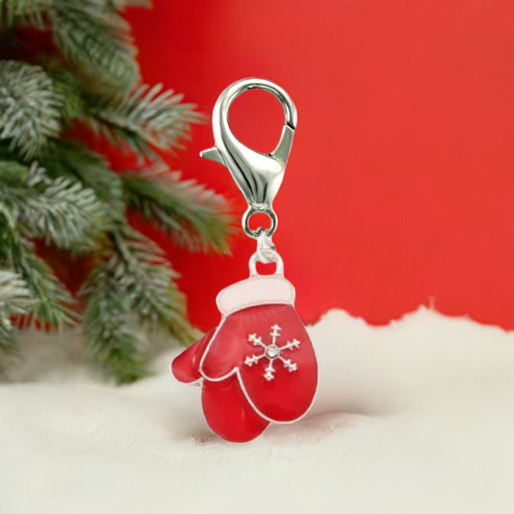 A red mitten-shaped charm with a silver lobster clasp, decorated with a white snowflake on the front, set against a festive background with red and white faux snow and green pine branches.