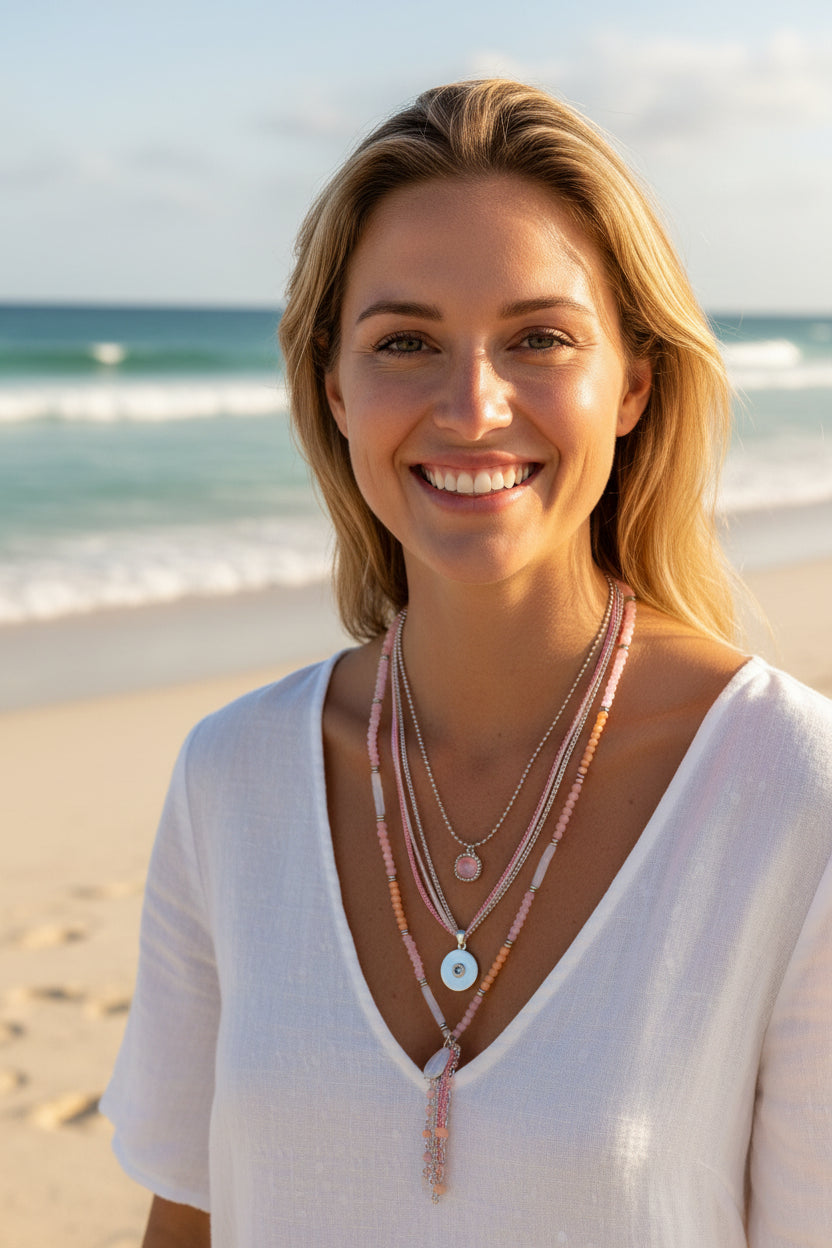 Woman wearing a white off-shoulder top with layered necklaces on a beach.