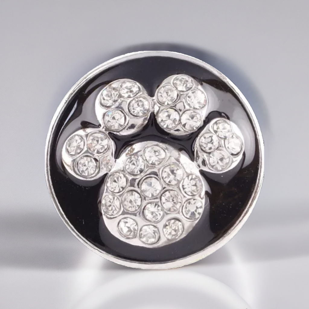 A round silver-toned snap button featuring a paw print design paved with clear rhinestones on a glossy black background.