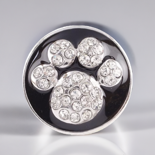 A round silver-toned snap button featuring a paw print design paved with clear rhinestones on a glossy black background.