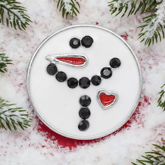 A round white snap button featuring a snowman face design with black dots, a red carrot nose, and a red heart, photographed on a snowy white surface with pine branches around the edges.