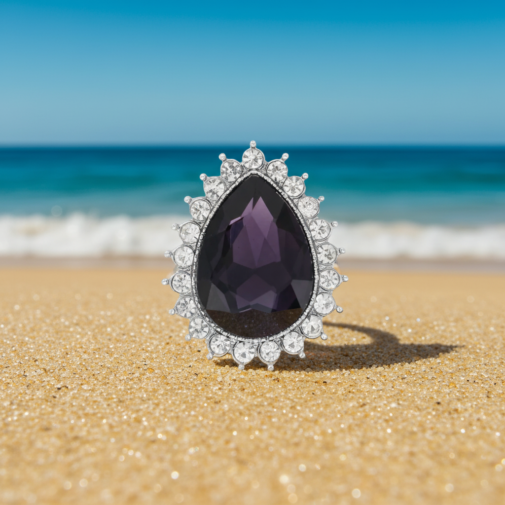 A teardrop-shaped silver snap featuring a faceted dark purple center stone surrounded by a halo of clear round stones, set against a blurred beach background.