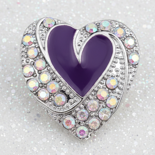 A heart-shaped silver statement snap with a deep purple enamel center and a border of iridescent rhinestones, resting on a sparkling silver surface.