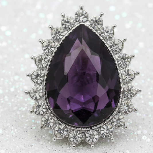 A teardrop-shaped purple gemstone snap set in silver-toned metal with surrounding clear pavé stones, photographed on a sparkly white background.