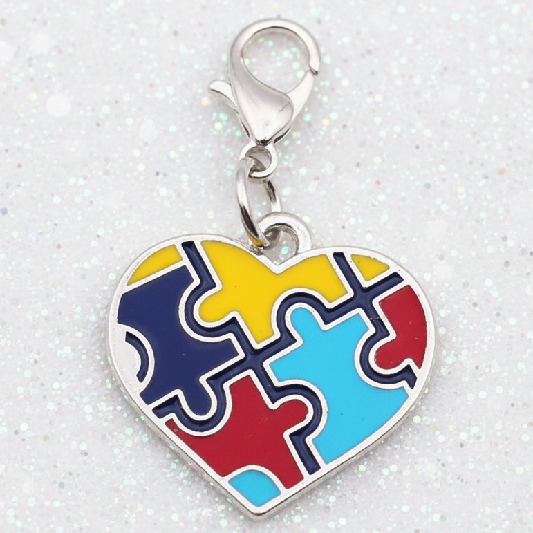 A silver heart-shaped charm with four interlocking puzzle pieces in yellow, blue, red, and light blue, attached to a silver lobster clasp, set against a pink and white background.