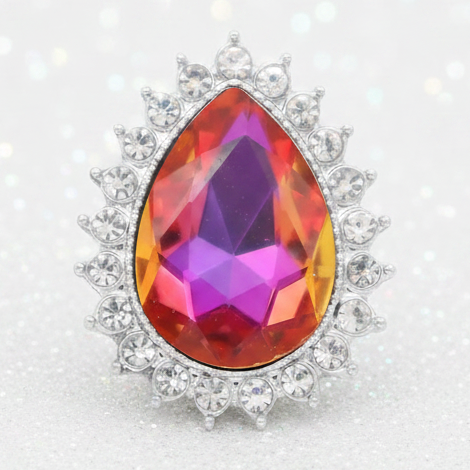 A teardrop-shaped gemstone snap in a silver halo setting, featuring a faceted crystal with a vibrant iridescent color shift from purple and pink to orange and yellow, surrounded by small clear stones on a sparkling white background.