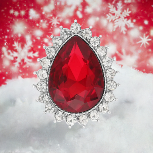A teardrop-shaped red gemstone snap button with a silver-toned halo of clear rhinestones, set against a festive red and white snowy background with snowflake graphics.