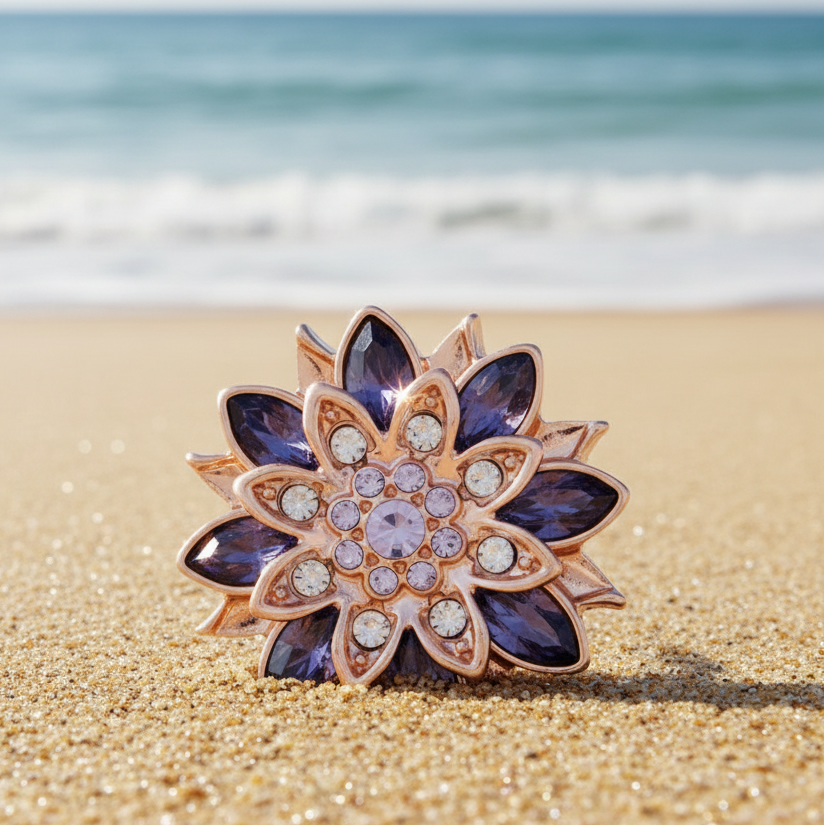 A rose gold flower-shaped snap button with deep purple gemstone petals and a central cluster of clear and light purple rhinestones, displayed on a sunny beach