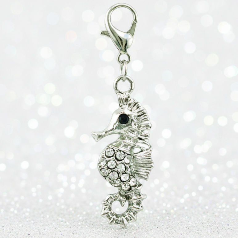 A silver-toned seahorse charm embellished with clear rhinestones and a small black eye, attached to a lobster clasp, set against a sparkling white bokeh background.