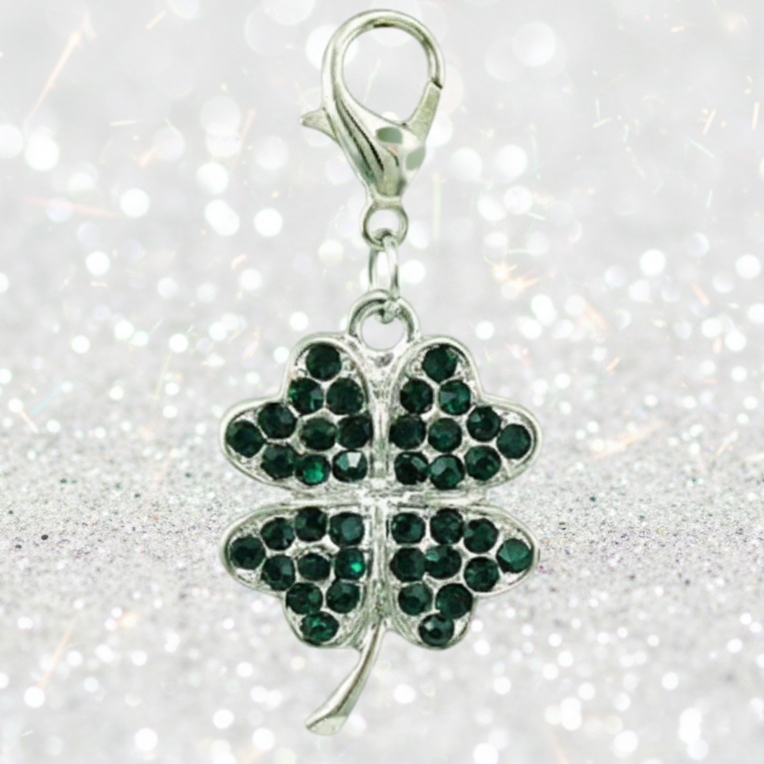 A silver shamrock charm embellished with dark green rhinestones, attached to a silver lobster clasp, set against a sparkling white glitter background.