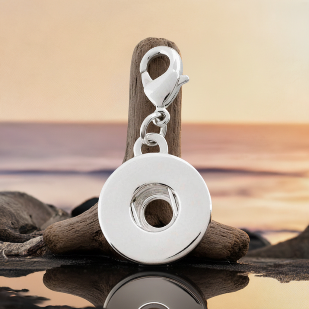 A polished silver metal round keyring or charm with a lobster clasp, displayed on a piece of driftwood over a beach sunset background.