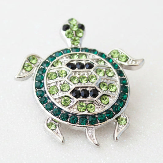 A turtle-shaped snap button featuring a silver-tone metal frame set with green and black rhinestones, photographed on a white background.