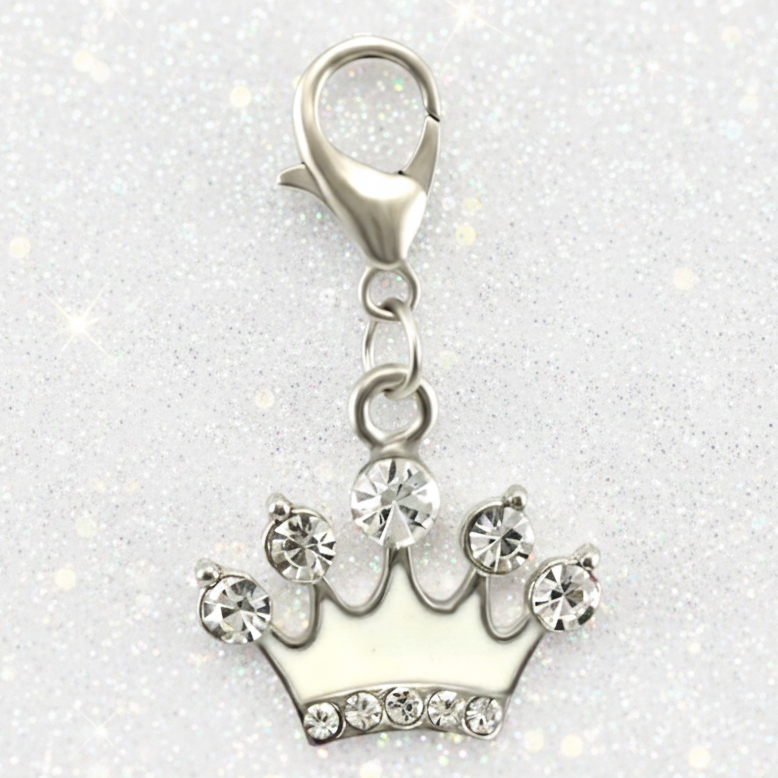 A silver crown-shaped charm with clear rhinestones and a lobster clasp, displayed in front of pink gift boxes on a purple surface with scattered confetti.