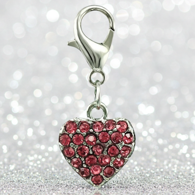 A silver heart charm embellished with small pinkish-red rhinestones, attached to a silver lobster clasp, set against a sparkling silver glitter background.