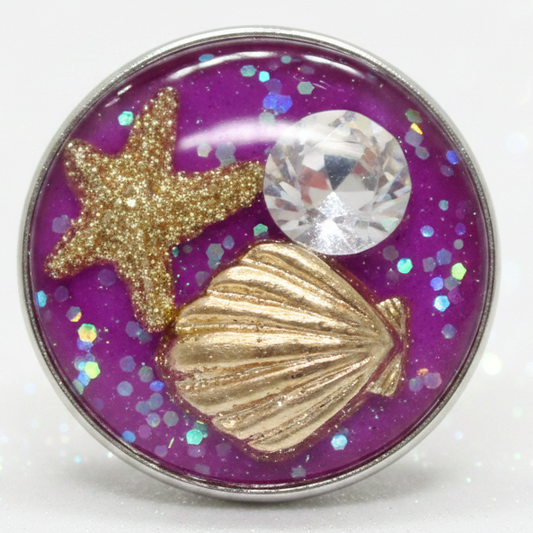A round snap button featuring a glittery purple background with a gold starfish, a gold seashell, and a clear faceted rhinestone, set in a silver-toned metal rim.