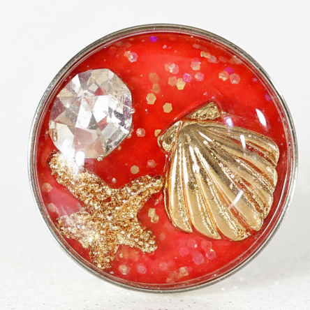 A round resin snap button featuring a vibrant red background filled with glitter, a faceted clear crystal, a gold seashell, and a gold starfish, displayed on a clean white surface.