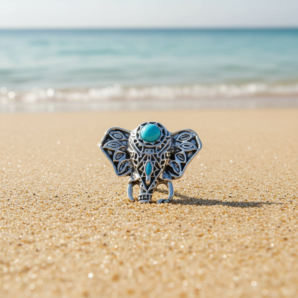 An ornate silver elephant snap button with turquoise blue gemstone accents, displayed on a beach