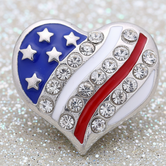A heart-shaped American flag statement snap with silver-tone metal, white enamel, a blue canton with silver stars, and a red stripe, embellished with clear rhinestones, set against a glittery silver background.