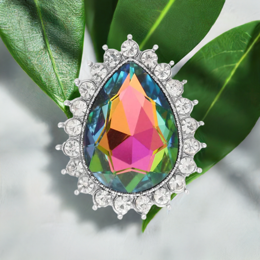 A teardrop-shaped iridescent gemstone statement snap with a silver-tone halo of clear crystals, displayed on green leaves against a light gray background.