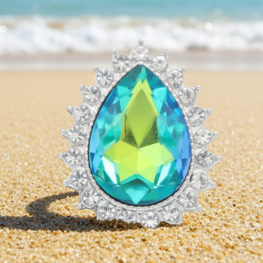 A teardrop-shaped turquoise and lime green gemstone snap with a silver sunburst halo, displayed on a stone surface with a blurred sunset background.