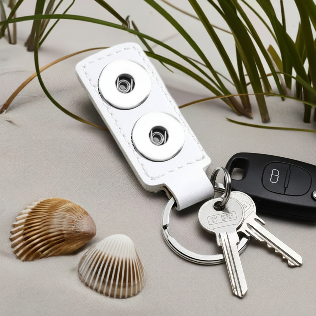 A white faux-leather keychain wallet with two silver metal snap buttons and attached keys, styled on a light beige surface with seashells and green grass.