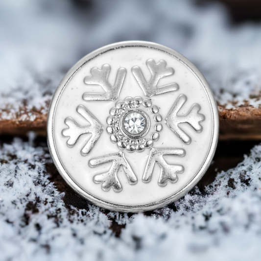 A round silver-toned snowflake snap button with a clear central gemstone, photographed close up against a snowy, frosty background.