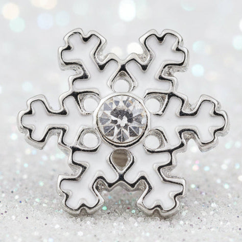 A silver snowflake-shaped snap button with a clear central gemstone, displayed on a smooth white stone with a soft, snowy, blurred background.